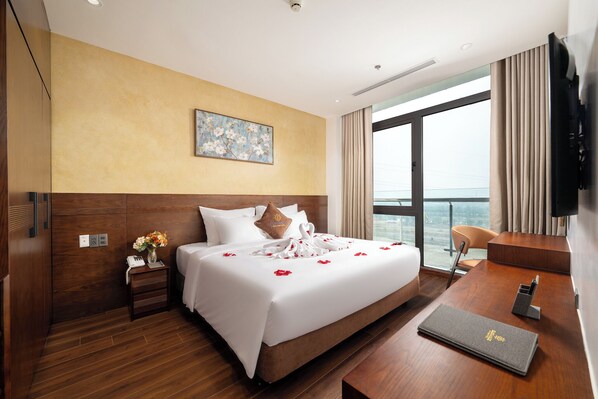 A hotel room with a large bed, a wooden headboard, a painting on the wall, a desk with a chair, and a view of the city through the window.