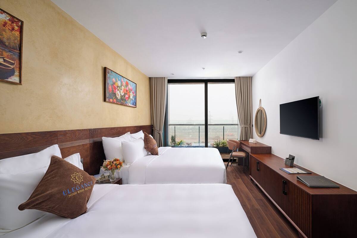 A hotel room with two beds, a desk, a TV, and a large window with a view.