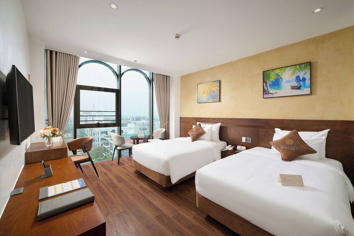 A hotel room with two beds, a desk, a chair, and a large window with a city view.