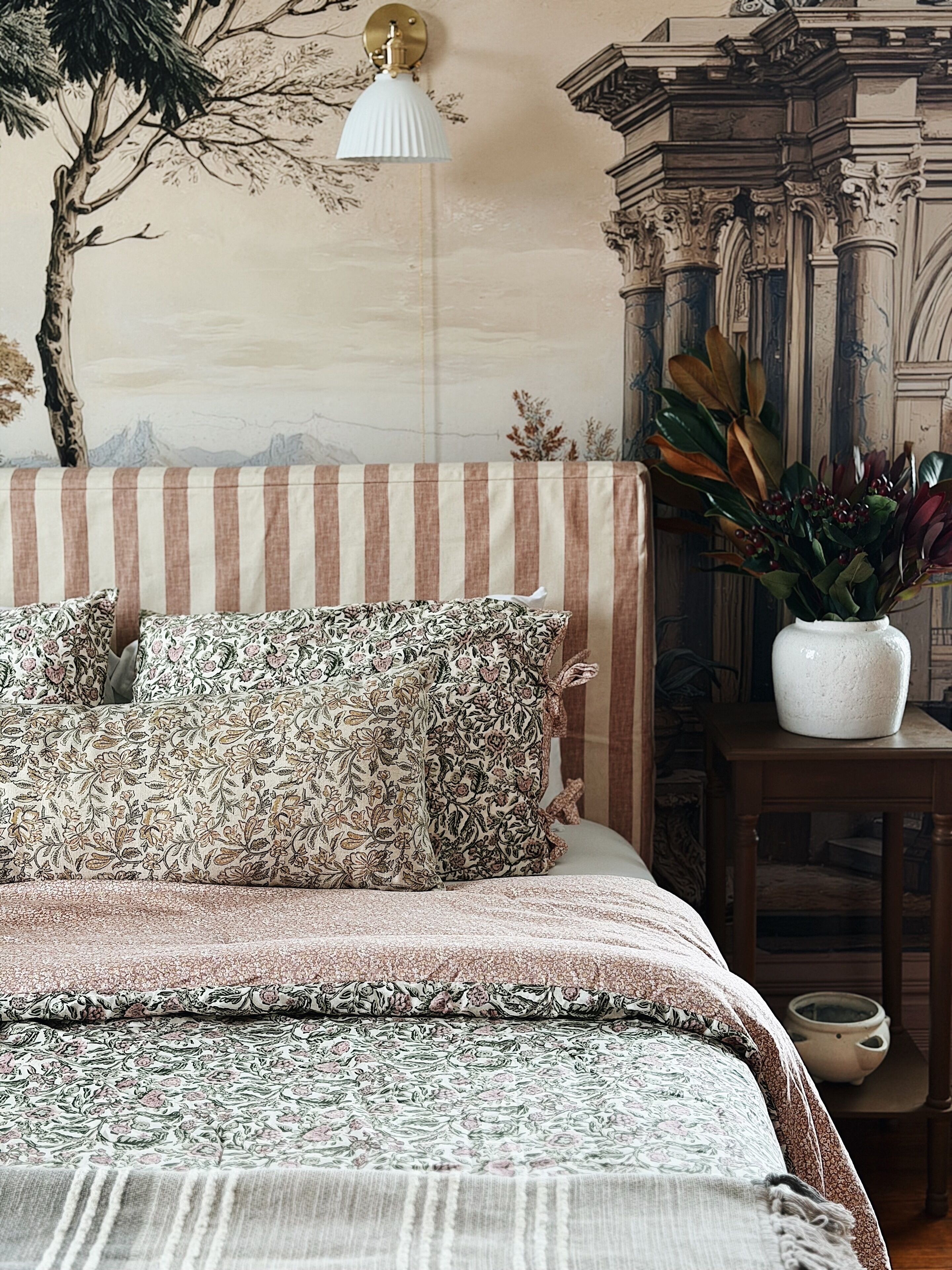 A bed with floral-patterned pillows and a striped headboard, a vase with a plant, and a wall with a mural.