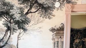 Un aparador de madera antiguo con cajones y un espejo, ubicado junto a una pared con un mural de un árbol y detalles arquitectónicos.