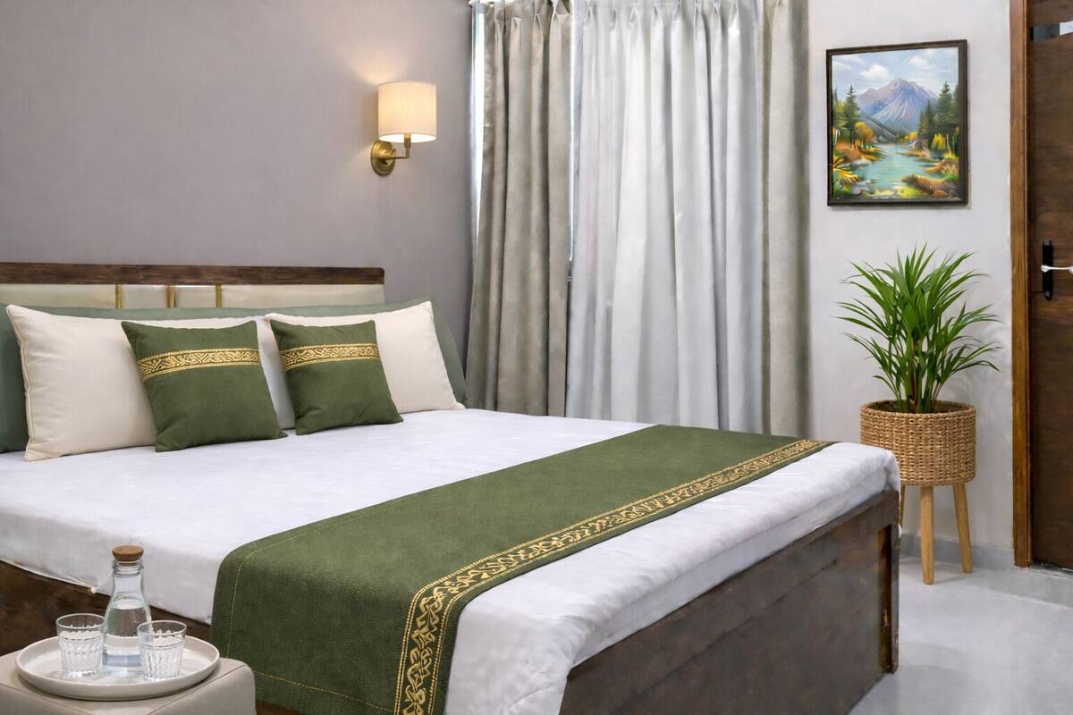 Una cama bien hecha con colcha verde y blanca, cabecera de madera y mesita de noche con una botella y vasos.