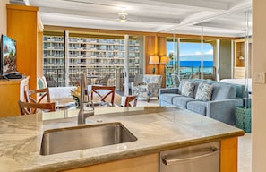 A modern kitchen with a sink, dining area with chairs and table, a sofa, and a view of a building with balconies. - Maui Resort Rentals: The Whaler 762 - 7th-Floor Studio, Stunning Pacific Ocean and Black Rock Views! (Lahaina)