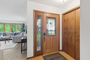 Eine Holztür mit Glasfenster, eine Fußmatte, ein Sofa und ein Fenster mit Blick auf Grünflächen.