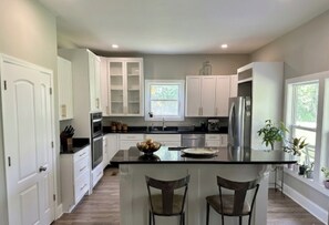 A modern kitchen with white cabinets, a central island, bar stools, and a window providing natural light. - Terrific 4-bedroom house in charming West Point (West Point)