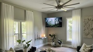 A living room with a ceiling fan, a flat-screen TV, a glass coffee table, and a sofa. - Terrific 4-bedroom house in charming West Point (West Point)
