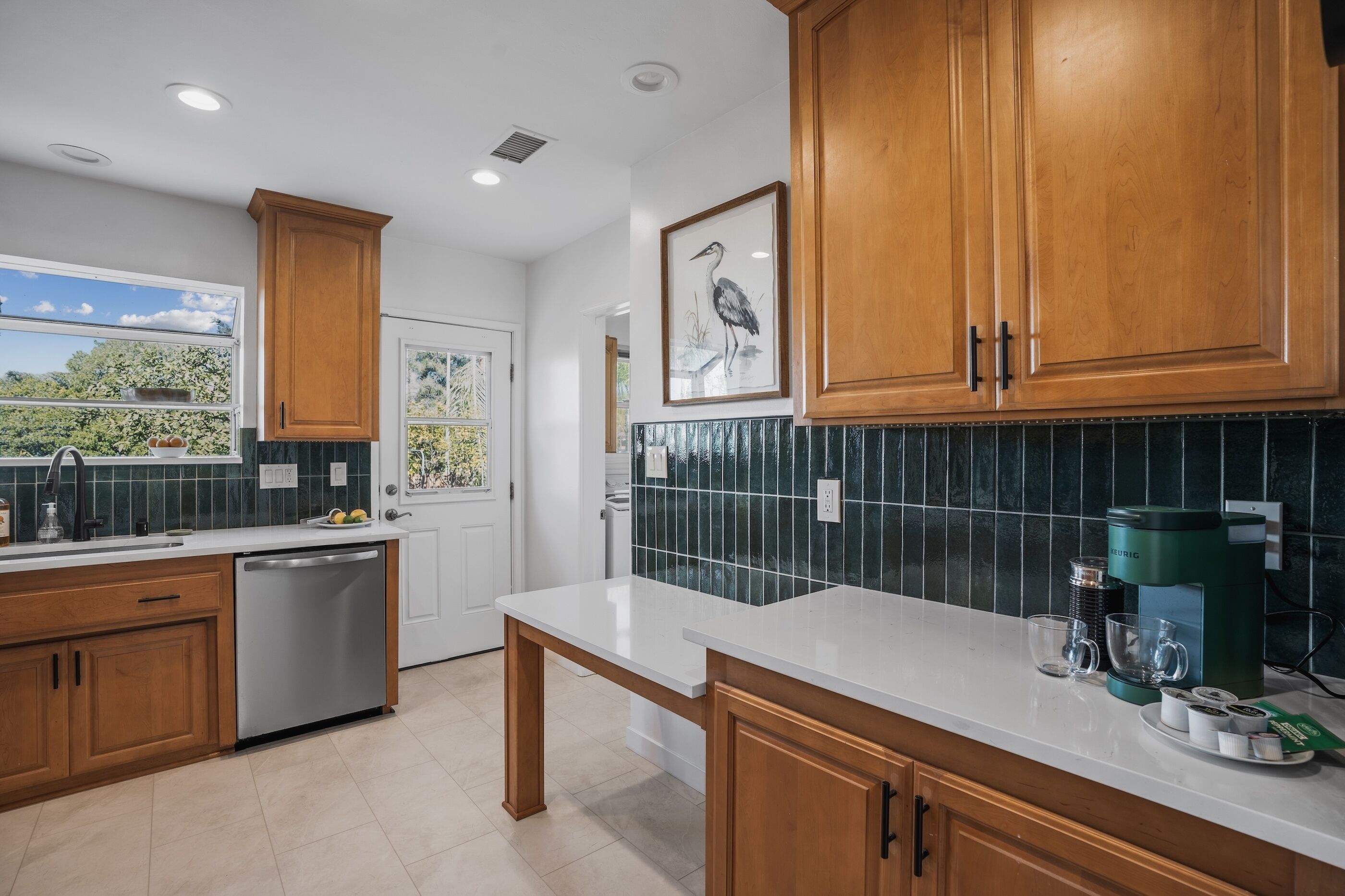 A modern kitchen with wooden cabinets, a white countertop, a green coffee maker, and a window with a view of greenery.