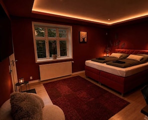 A bedroom with a bed, a window, a radiator, a red rug, and a small table with a lamp. - Crimson Autumn - Nordic Suite (Aalborg)