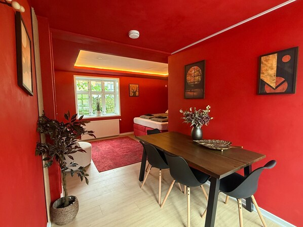 A red-walled room with a wooden dining table, black chairs, a radiator, a window with a view, and a plant. - Crimson Autumn - Nordic Suite (Aalborg)