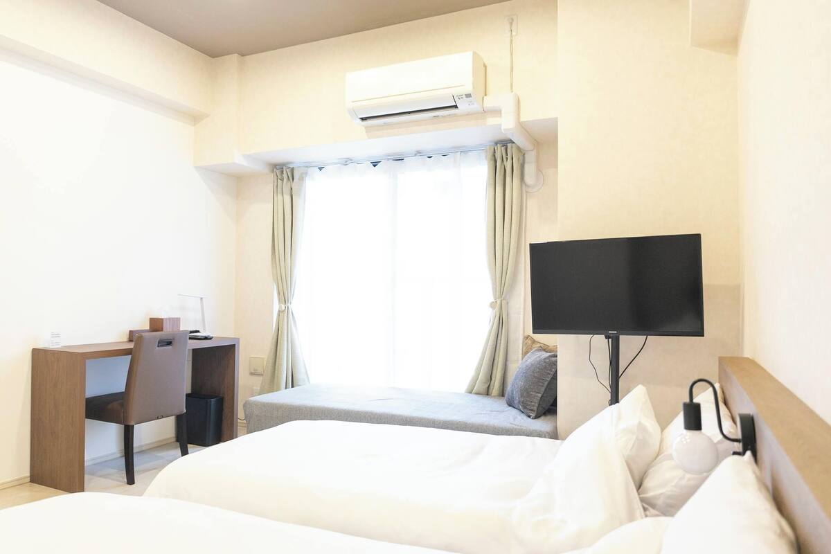 A hotel room with a bed, a desk, a chair, a TV, and an air conditioning unit.