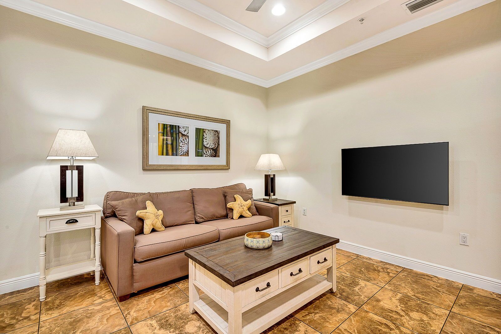 Un soggiorno con un divano beige, due cuscini a forma di stella marina, un tavolino in legno, un quadro incorniciato, una televisione a schermo piatto e un comodino con lampade.