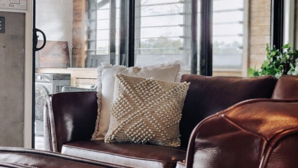 Un canapé en cuir agrémenté d’un coussin tissé, dans une pièce dotée de grandes fenêtres donnant sur une zone industrielle.