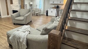 A living room with a grey sofa set, a coffee table, and a fireplace. There is a dining area with a table and chairs, and a kitchen with cabinets and a stove. A wooden staircase is visible on the right. - Relaxing Farmhouse in beautiful Butler (Butler)