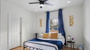 Une chambre à coucher avec un lit, un ventilateur de plafond, une fenêtre avec des rideaux et une petite table avec une lampe.