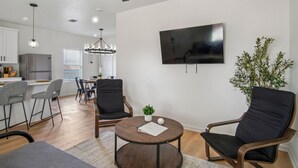 Un salon moderne avec une table basse ronde, deux fauteuils noirs, un coin repas agrémenté d’un lustre, et une cuisine en arrière-plan.