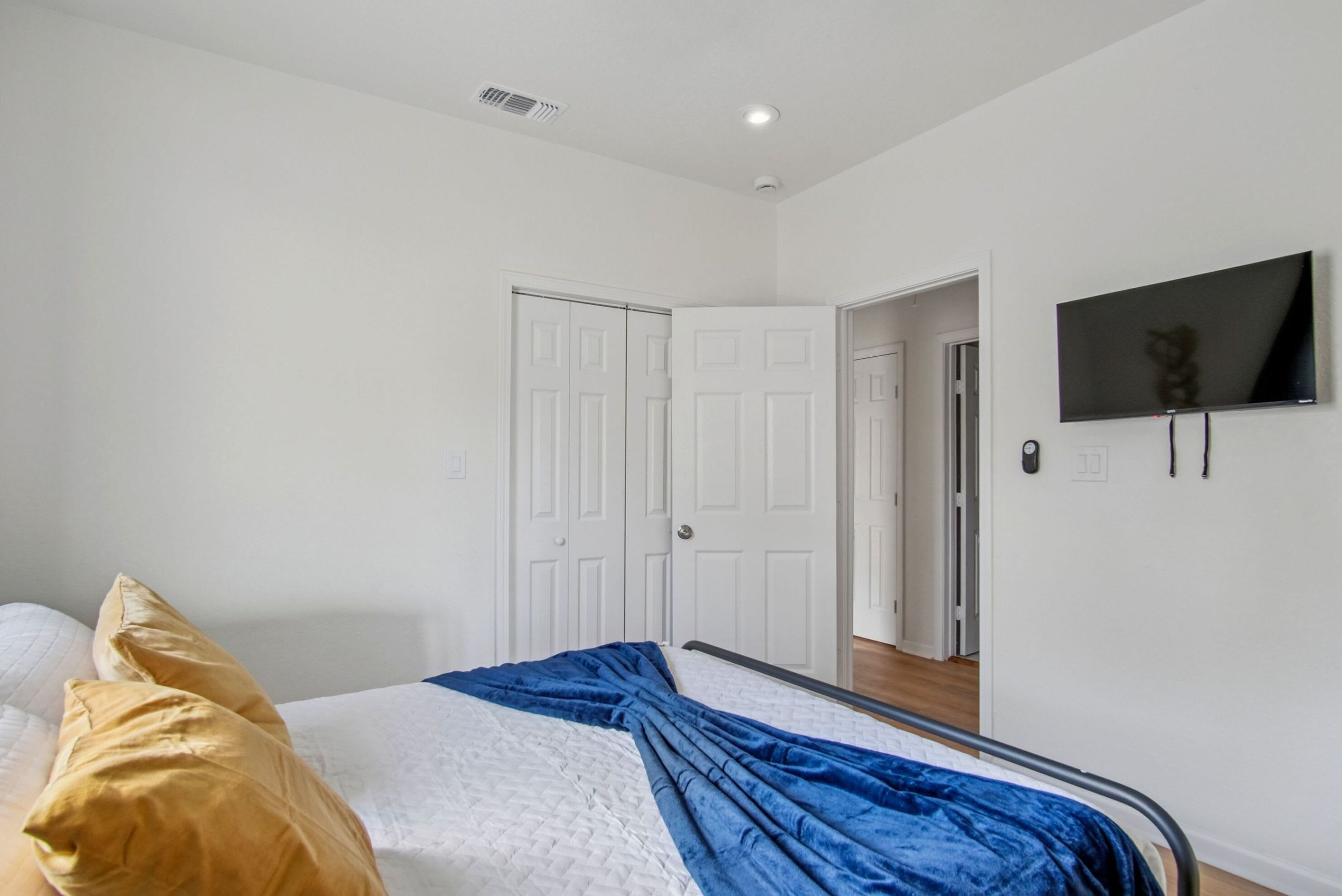 A bedroom with a bed, white walls, a TV mounted on the wall, and a door leading to another room.