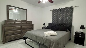 A bedroom with a bed, a dresser, a ceiling fan, a mirror, a nightstand, and a window with curtains. - Spacious Home in lovely Lehigh Acres (Lehigh Acres)