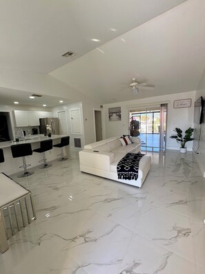 Uma sala de estar ampla com sofá branco, cozinha com armários brancos e espaço de jantar com banquetas.