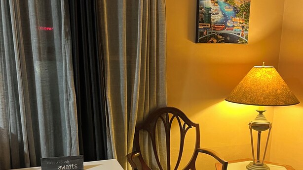 Una habitación de hotel con chimenea, una silla de madera con cojín floral, una mesita de noche con lámpara y un cuadro en la pared.