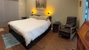 Ett sovrum med en stor säng, två sänglampor, ett trägolv, en mörk läderfåtölj och ett litet bord med en näsdukslåda.