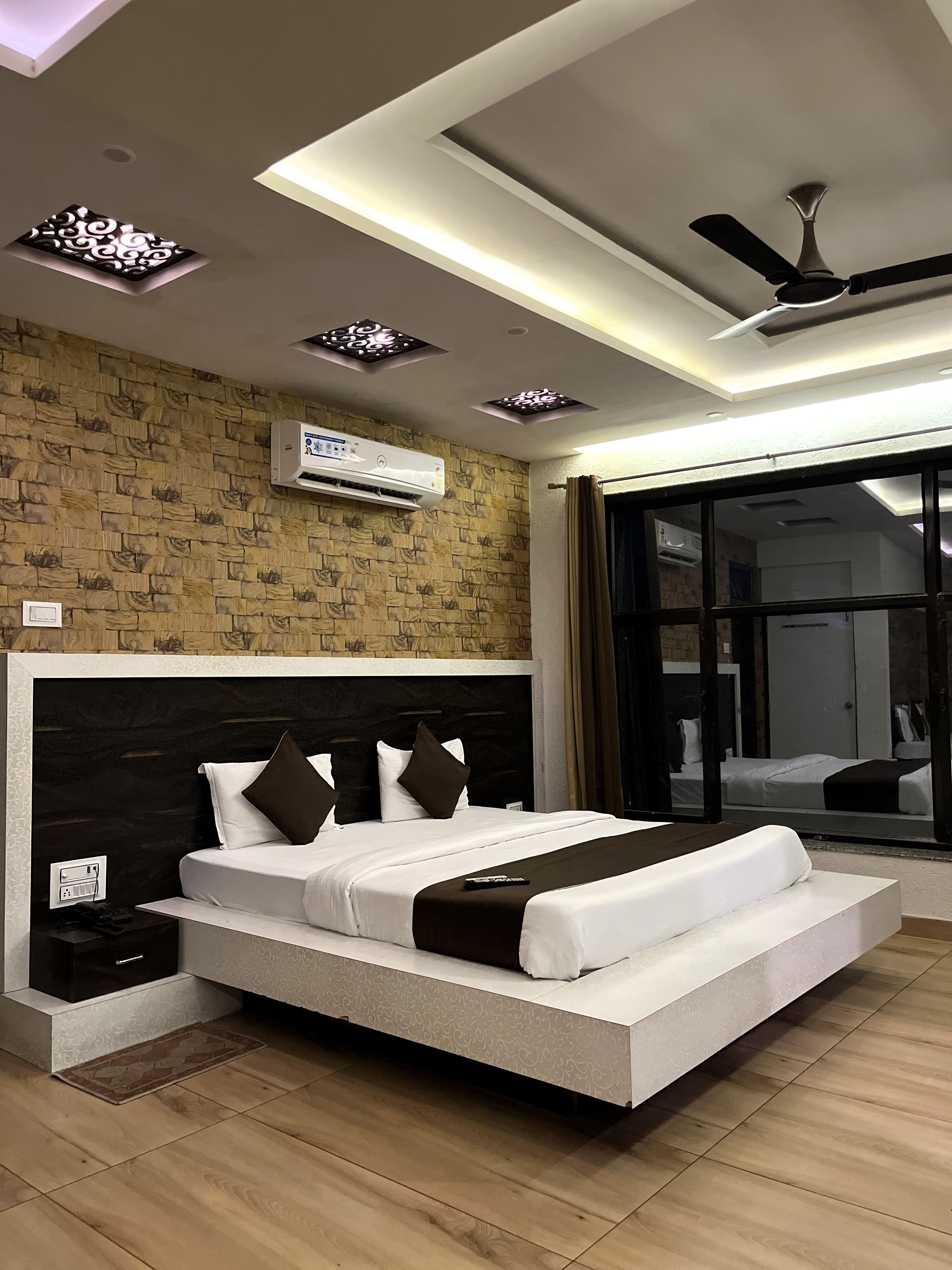 A modern hotel room with a large bed, ceiling fan, air conditioning unit, and a view through a glass door.