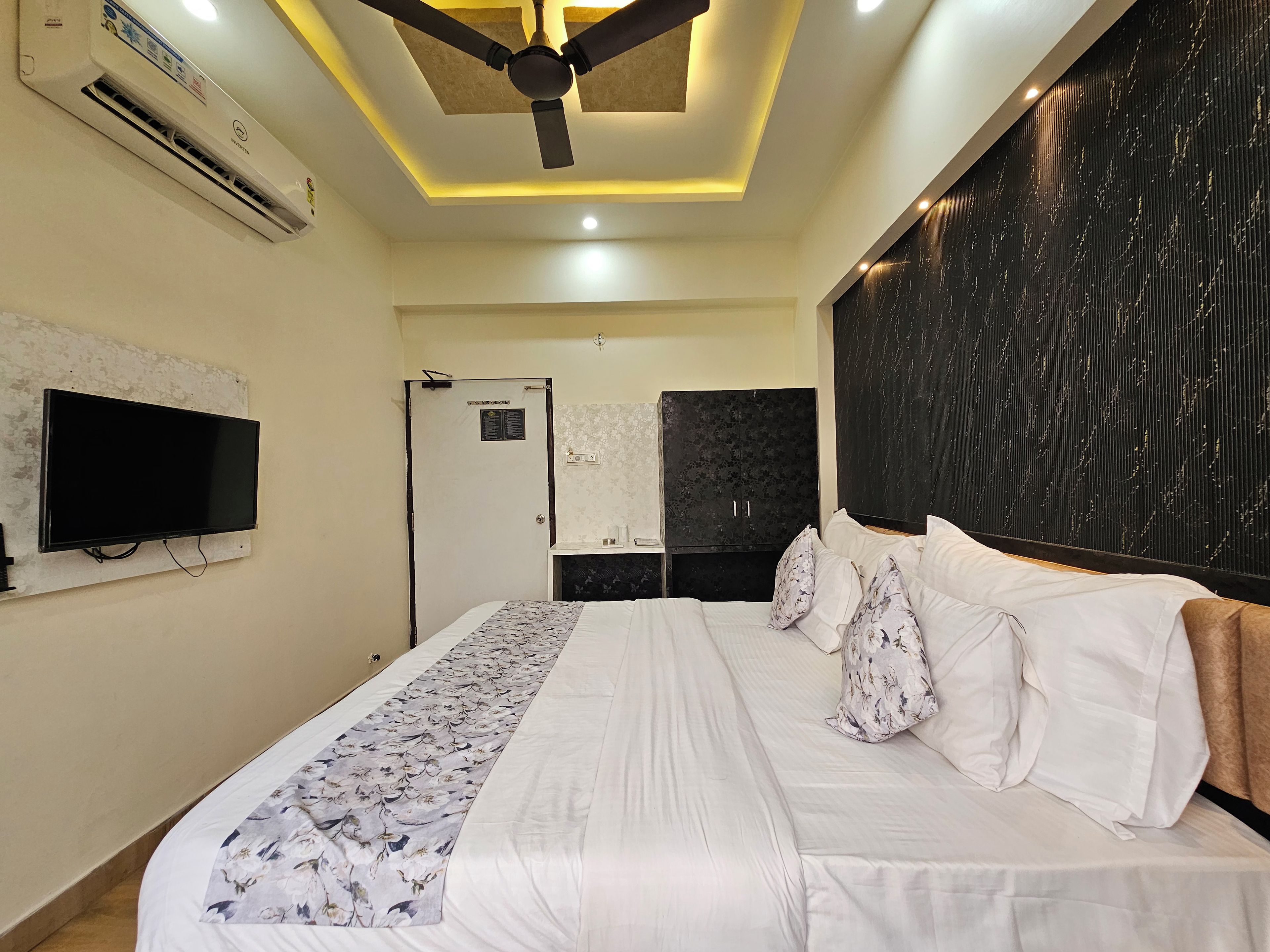 Una camera d'albergo con un letto, una TV a schermo piatto, un condizionatore d'aria, una ventola a soffitto e una testiera scura.