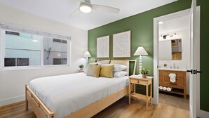 Un dormitorio con cama, mesitas de noche, ventilador de techo, ventana y un baño visible a través de una puerta abierta.