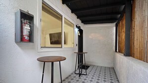 Pequeña zona de balcón cerrada, con suelo de baldosas, dos mesas, un extintor y una ventana.