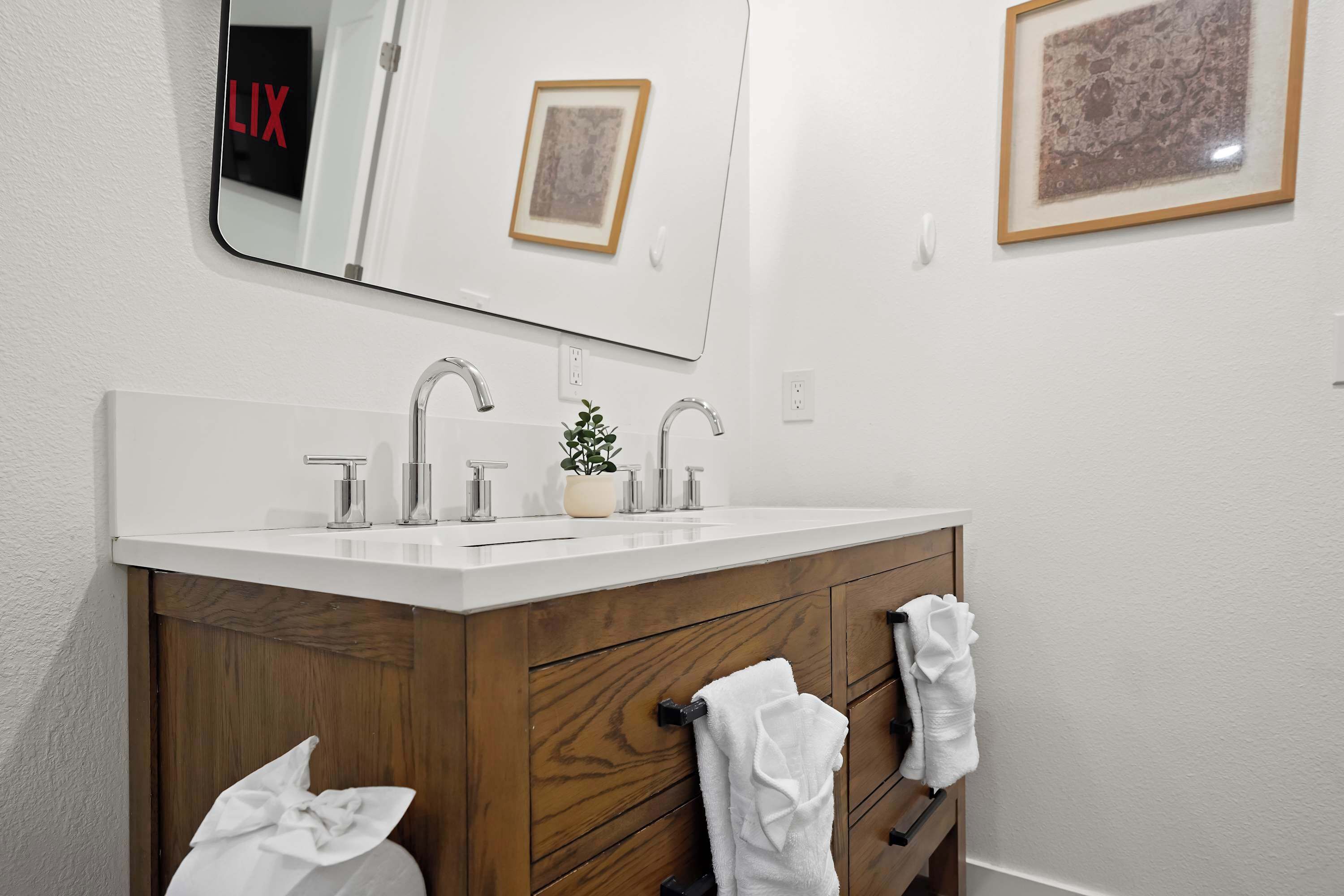 Ein Badezimmer mit einem weißen Waschbecken, zwei Wasserhähnen, einem Spiegel und einer Holz-Waschkommode mit zwei aufgehängten Handtüchern.