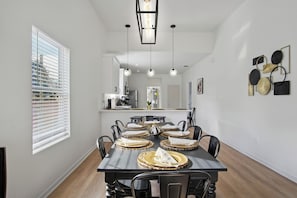 Un comedor con una mesa larga dispuesta para seis personas, una cocina al fondo y un amplio ventanal con persianas.