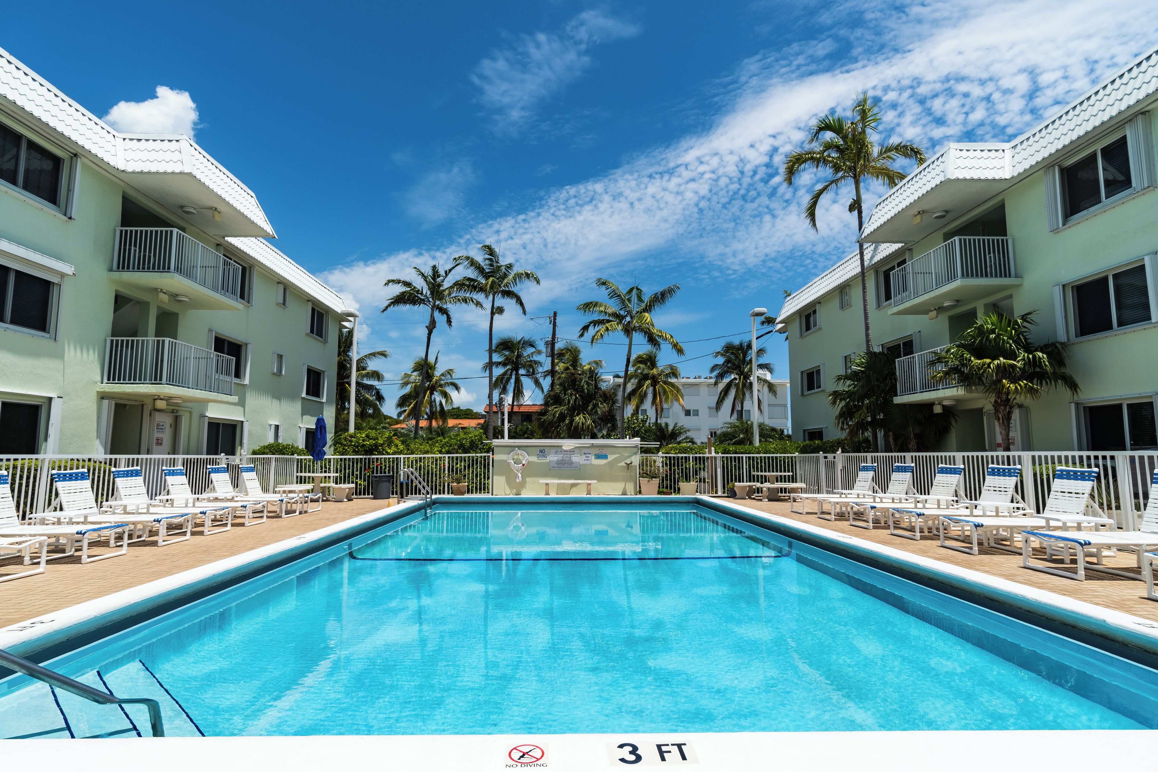 Ein Swimmingpool mit Liegestühlen, umgeben von einem weißen Zaun und Palmen im Hintergrund.