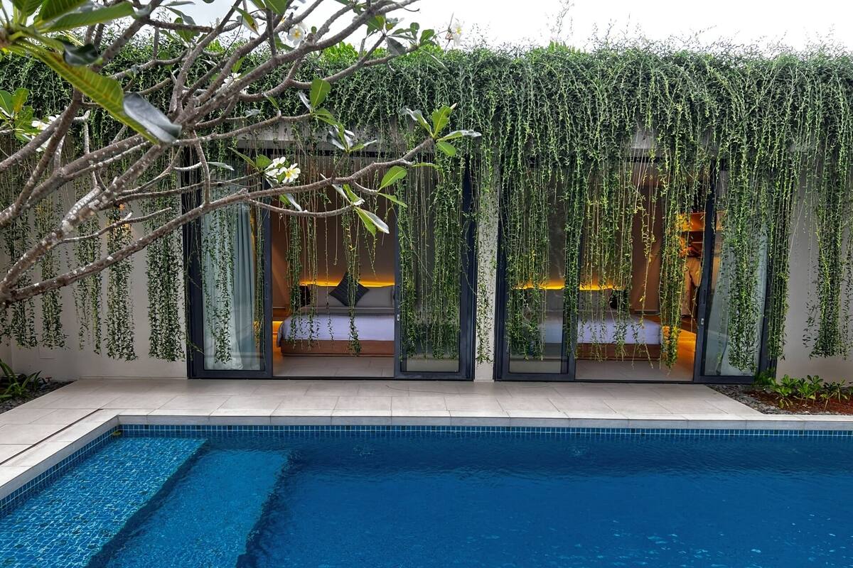 A modern poolside area with a clear view of a bedroom through glass doors, a swimming pool, and lush greenery.