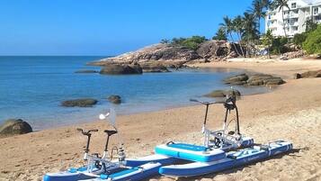 Une plage avec des kayaks et une planche de paddle, un ciel bleu dégagé et un bâtiment en arrière-plan.