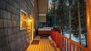 Una terrazza in legno con vasca idromassaggio e ringhiera in legno, affacciata su una foresta innevata.