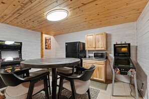 Une cuisine compacte avec une table ronde, deux chaises, des meubles en bois, un réfrigérateur noir, un four à micro-ondes et deux bornes d’arcade.
