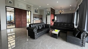 A modern living room with black leather sofas, a kitchen in the background, and a large sliding glass door leading to an outdoor area. - Anemos Resort By Tamastu (Cijeruk)