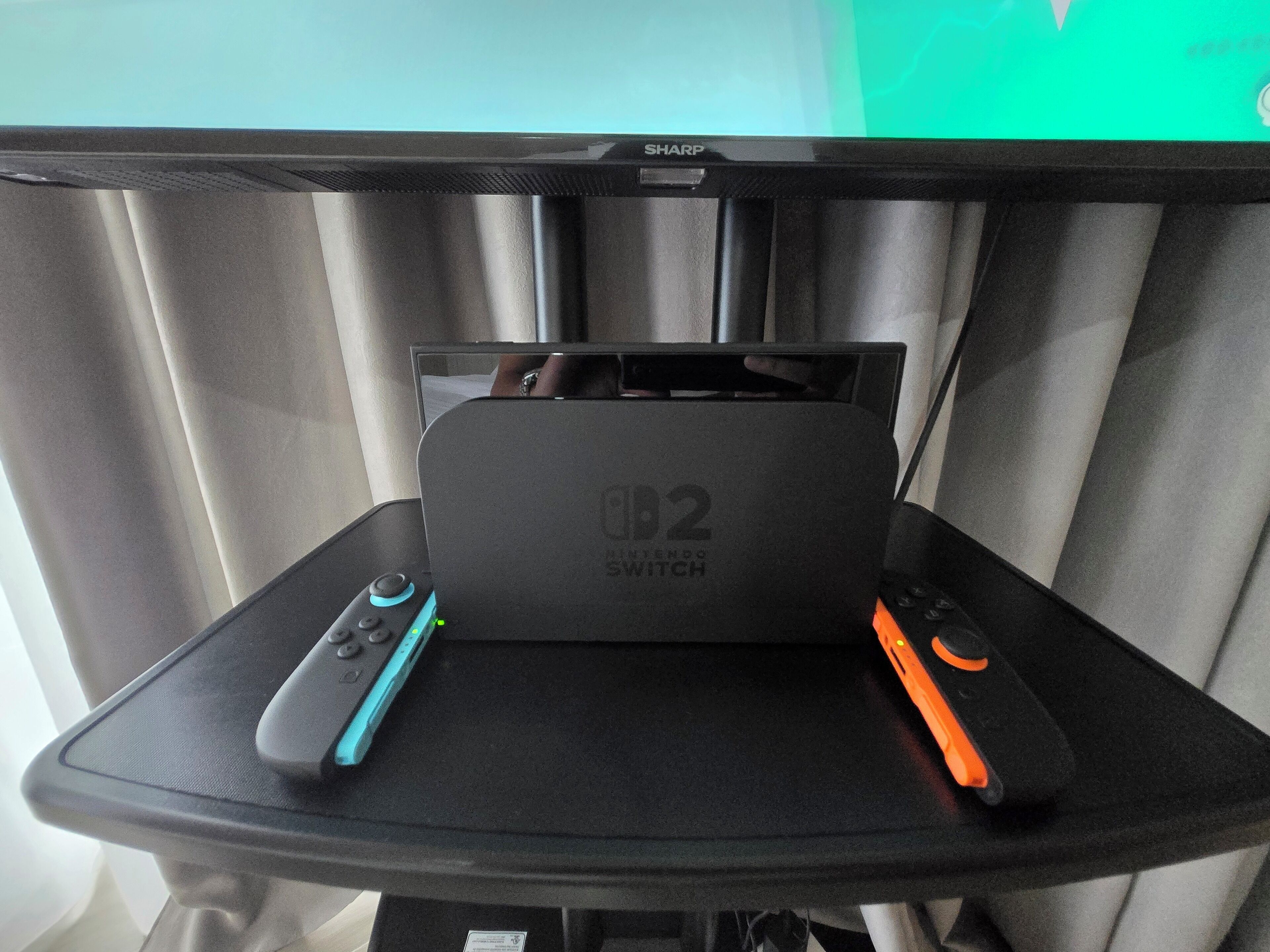 Ein schwarzer Tisch mit einem Nintendo Switch und zwei Controllern, einem blauen und einem orangen, vor einem Fernseher.