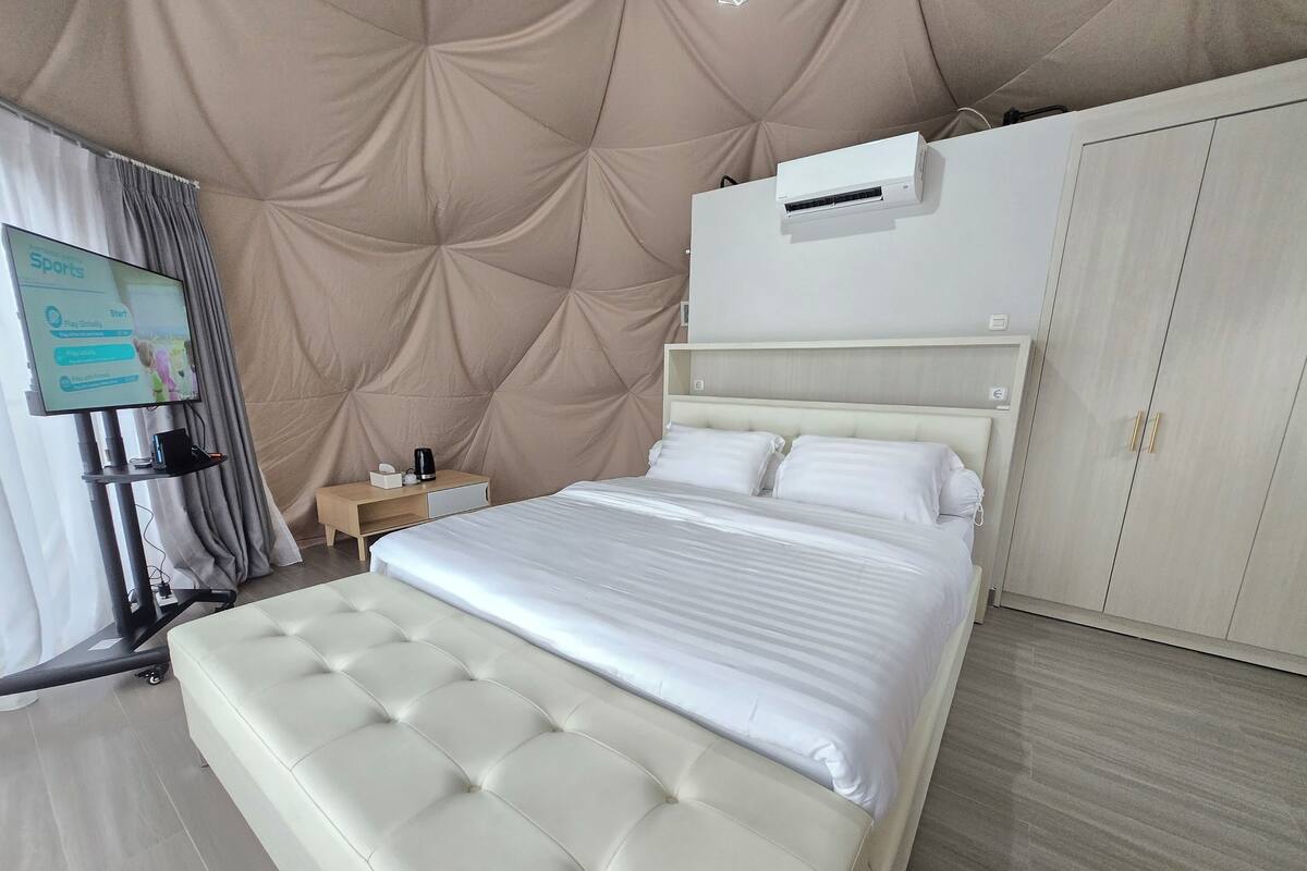 Une chambre d’hôtel moderne avec un grand lit, un téléviseur posé sur un meuble, une armoire et une petite table de chevet avec une lampe.