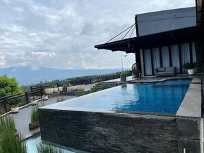 Una piscina infinita en la azotea con vistas a montañas, un área de estar moderna al aire libre y una planta grande en maceta.