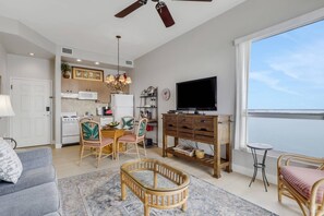 A modern living room with a grey sofa, wicker chairs, a TV on a wooden stand, and a large window with a view of the sea. - Penthouse-Level Retreat | Sweeping Estero Bay Views & Beautiful Sunrises | Resort Pool & Hot Tub (Fort Myers Beach)