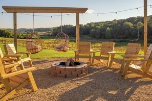 A wooden outdoor seating area with a fire pit and hanging chairs, overlooking a grassy landscape.