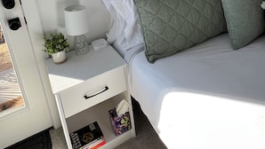 Un dormitorio ordenado con cama, almohadas, una mesita de noche con lámpara, una planta pequeña y una estantería con libros.