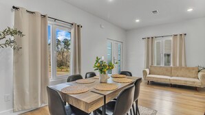 A dining area with a wooden table, black chairs, and a vase with flowers. There is a beige sofa in the background and a window with curtains. - Spacious new 3-bedroom house in  Spartanburg (Spartanburg)