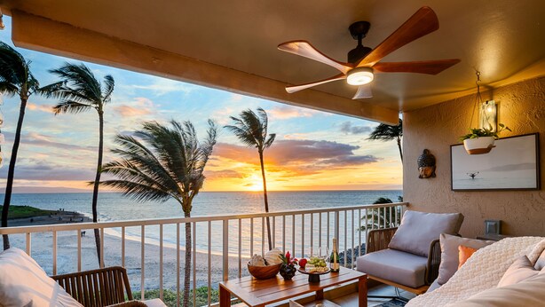 A balcony with wicker furniture, a wooden table with fruits and a bottle, and a view of the sunset over the ocean.