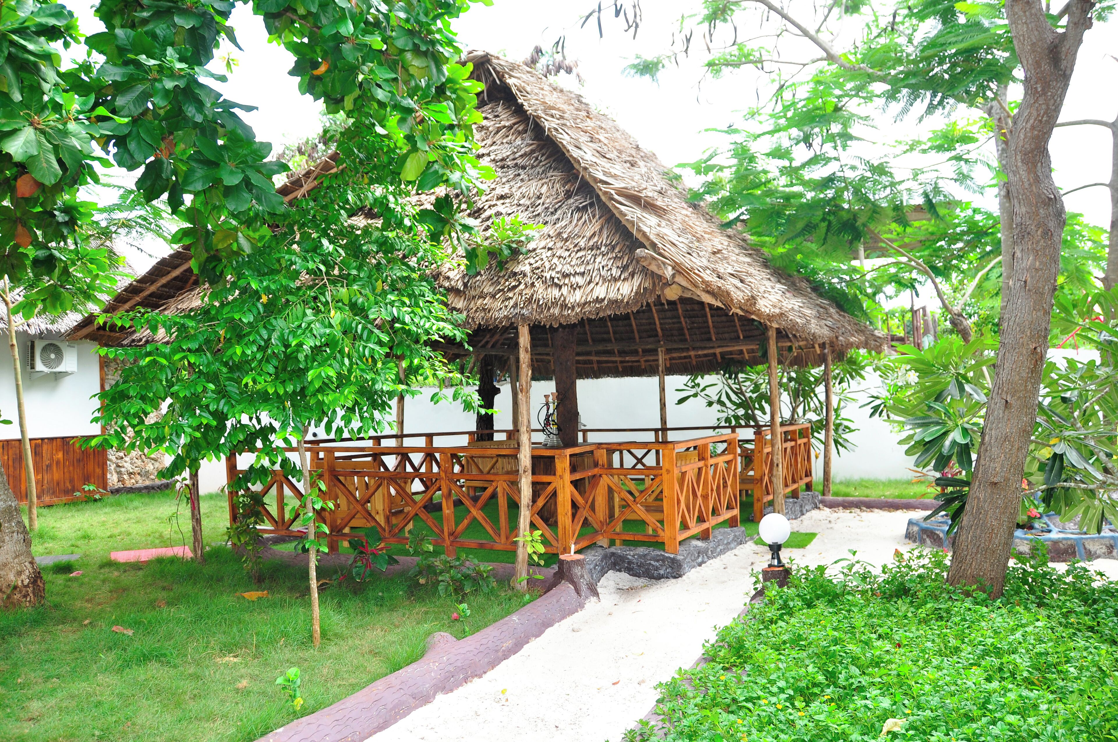 A thatched-roof gazebo with a wooden railing, surrounded by lush greenery and a paved pathway.