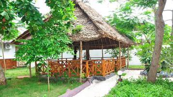 A thatched-roof gazebo with a wooden railing, surrounded by lush greenery and a paved pathway.