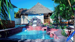 A pool area with a thatched-roof hut, surrounded by tropical plants and a rocky landscape. - Jodari bungalow kizimkazi Zanzibar (Kizimkazi)