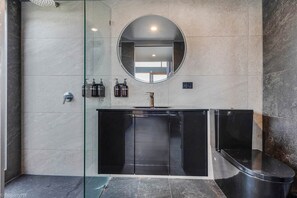 A modern bathroom with a glass shower enclosure, a black vanity with a sink, and a round mirror above the vanity. - Sanctuary Hobart CBD (Hobart)