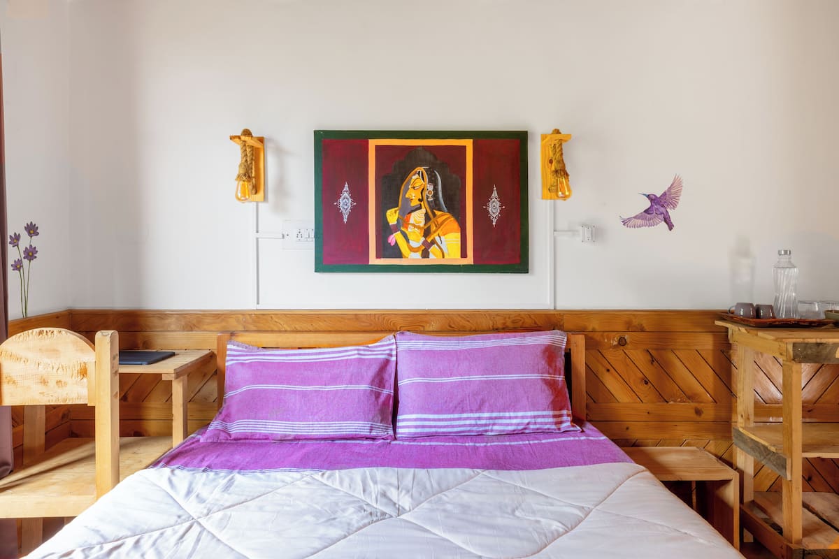 A bedroom with a wooden headboard, a bed with purple and white bedding, a small wooden table, and a wall-mounted artwork.