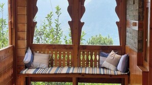 A wooden bench with cushions, a stone wall, and a view of greenery and mountains. - Huts And Hunters Cottage Manali (Kullu)